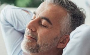 Older man laying on his back with his eyes closed and his hands behind his head